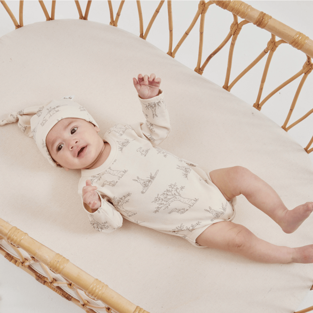 A baby wearing a printed onesie and an Aster & Oak Organic Bunny Luxe Rib Knot Hat made of GOTS-certified organic cotton lying in a woven bassinet.