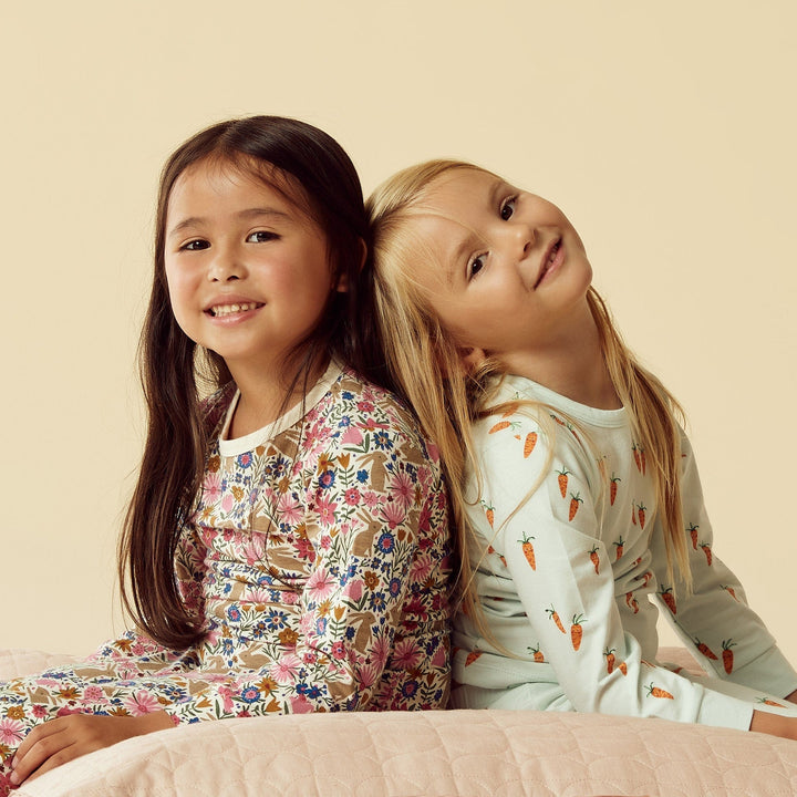 Two young girls smiling and sitting back-to-back against a plain background, wearing their Wilson & Frenchy Organic Long Sleeve Easter Pyjamas.