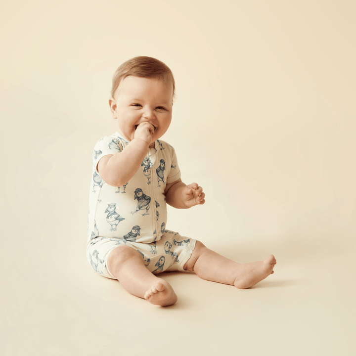 Baby sitting on a beige background, smiling with one hand in mouth, wearing a Wilson & Frenchy Organic Boyleg Zipsuit.
