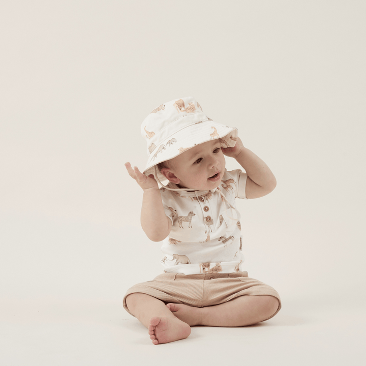 A baby sitting on the floor wearing an Aster & Oak Bucket Hat - LUCKY LASTS - MEADOW & LION ONLY and shorts.