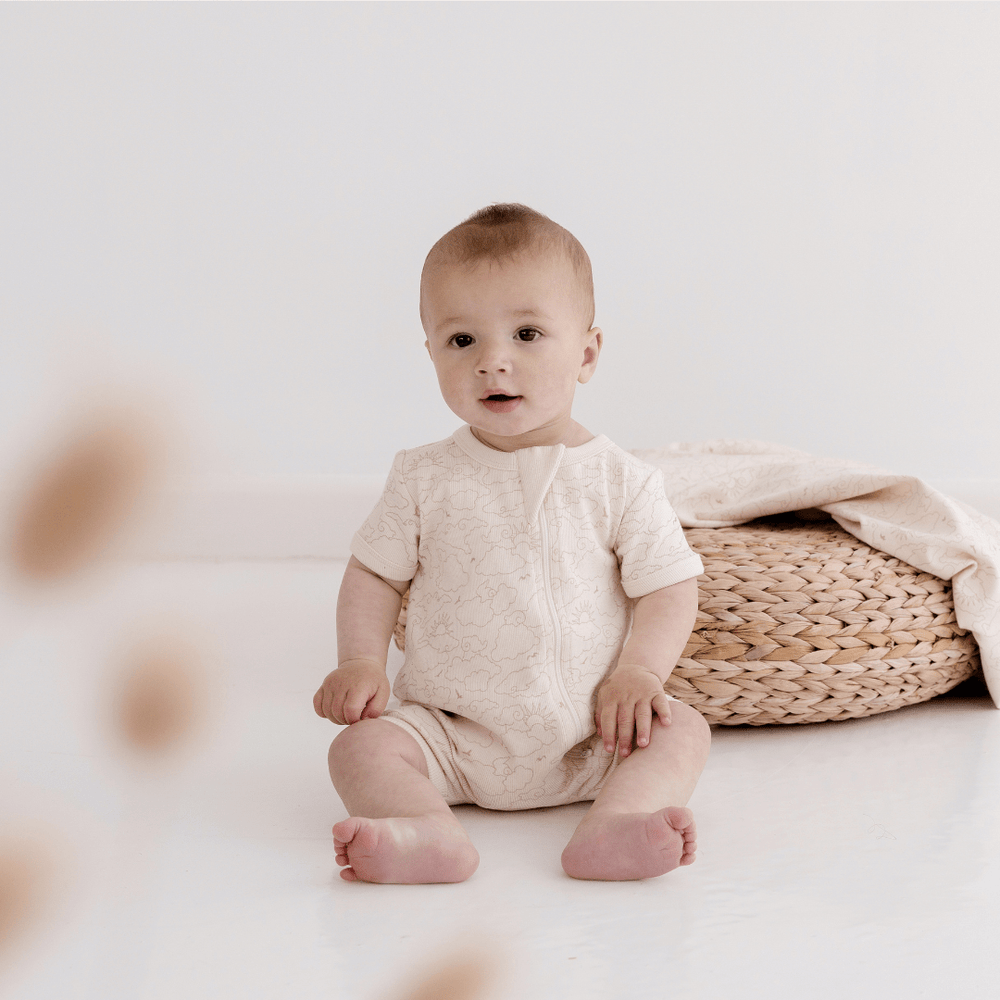 Baby Wearing a Ribbed, Cream Coloured Boyleg Zipsuit With Sun & Cloud Pattern Sitting In Front Of A Rattan Stool With A Blanket Sitting On It