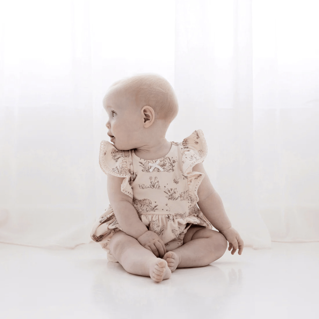 A baby in an Aster & Oak Organic Cotton Meadow Bubble Romper sits on the floor by the window.