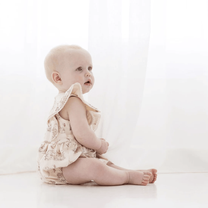 A baby in an Aster & Oak Organic Cotton Meadow Bubble Romper sits on the floor in front of a curtain.