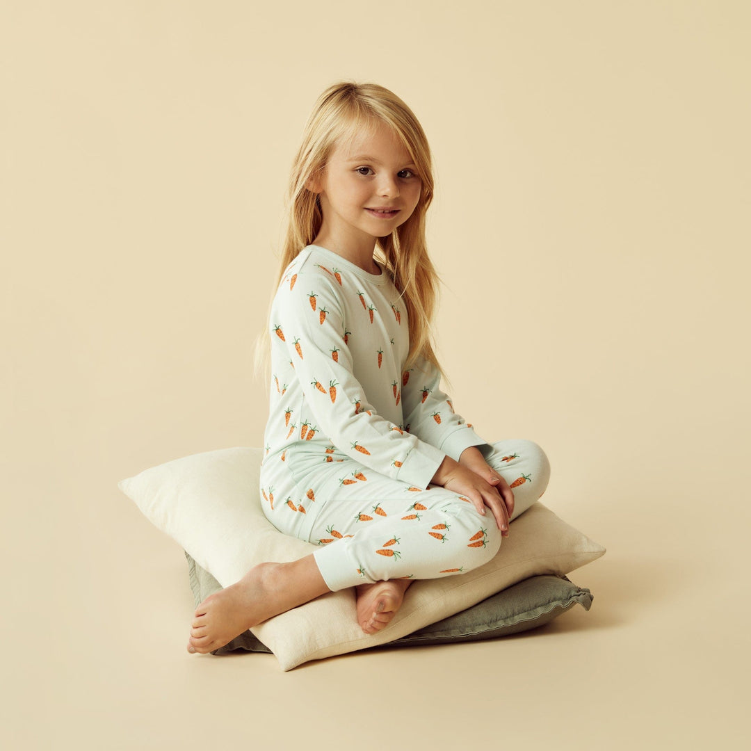 A child sitting on a stack of pillows wearing Wilson & Frenchy Organic Long Sleeve Easter Pyjamas against a beige background.