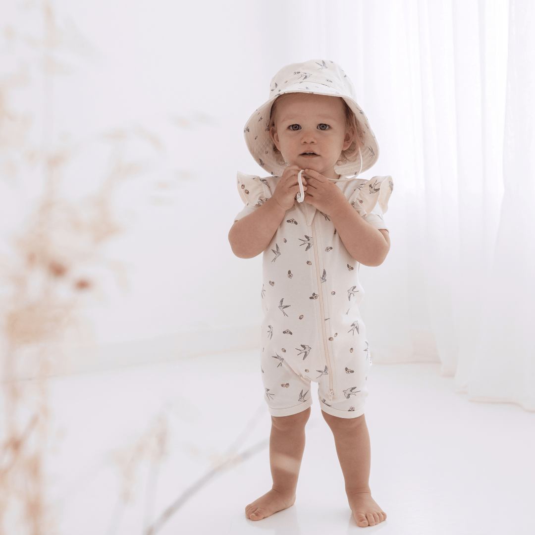 A baby wearing an Aster & Oak Bucket Hat - LUCKY LASTS - MEADOW & LION ONLY and shorts in a white room made of cotton canvas.