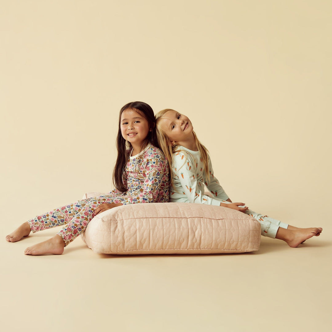 Two girls in Wilson & Frenchy Organic Long Sleeve Easter Pyjamas - LUCKY LASTS - 1 YEAR ONLY sitting back-to-back on a cushion against a beige background.