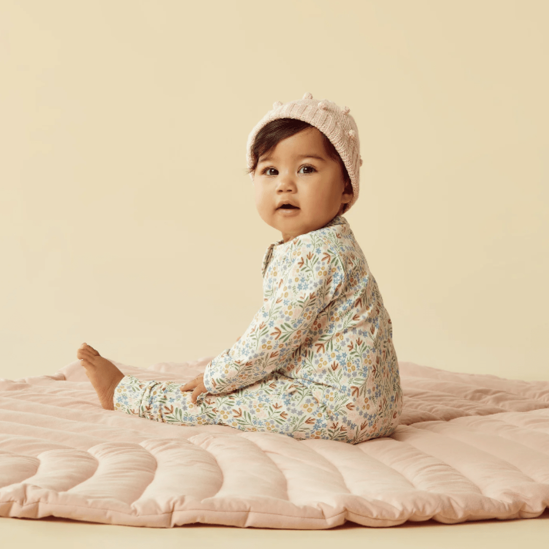 A baby wearing Wilson & Frenchy organic baby pyjamas and a knit hat sits on a soft pink surface against a pale yellow background.