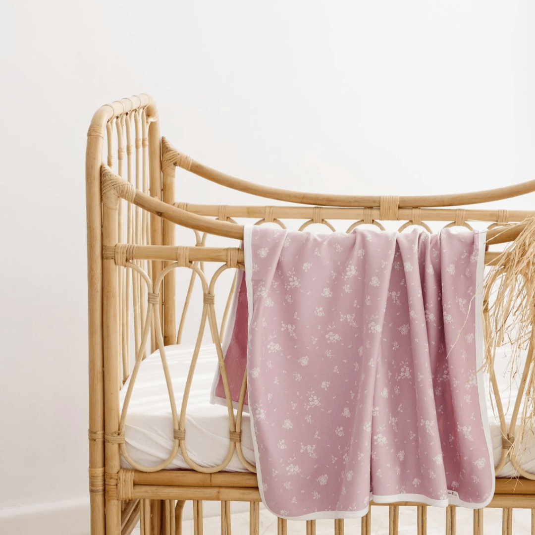 An Aster & Oak pink organic cotton baby swaddle wrap on a rattan crib.