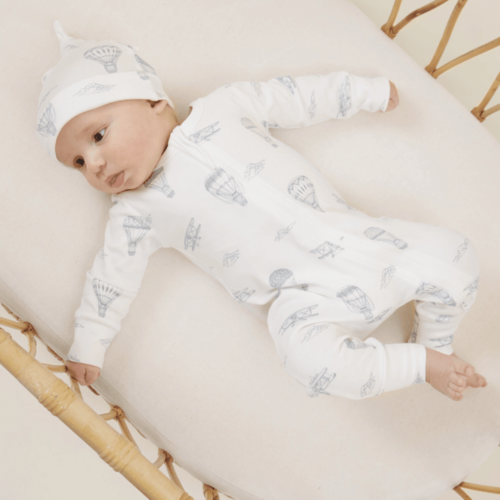 A baby wearing an Aster & Oak Organic Knot Baby Hat made of GOTS-certified organic cotton with hot air balloon prints lying on a cream-colored cushion.