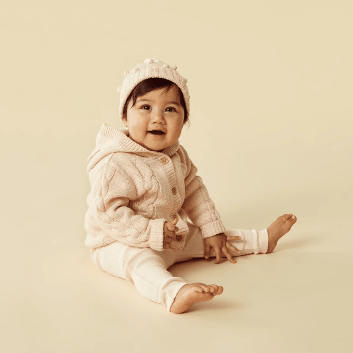 Baby in a pink Wilson & Frenchy Cable Knit Hooded Jacket and hat sitting against a beige background.