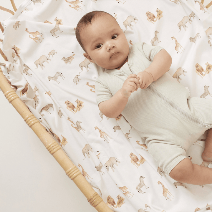 An Aster & Oak Organic Cotton Baby Swaddle Wrap, made of organic cotton, laying in a hand-illustrated print wicker basket with animals on it.