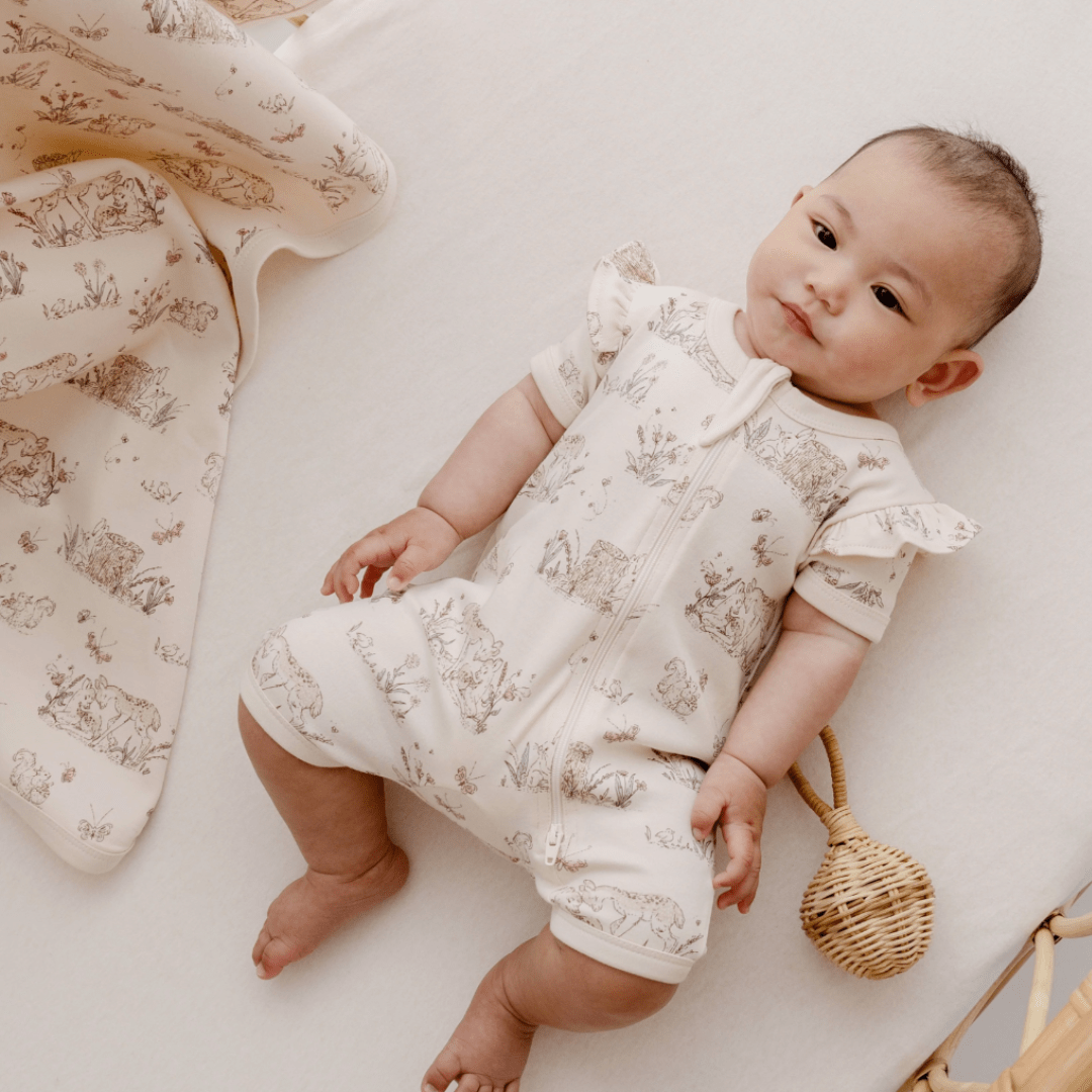 A baby is peacefully laying on a bed with an Aster & Oak Organic Cotton Flutter Sleeve Zip Romper.