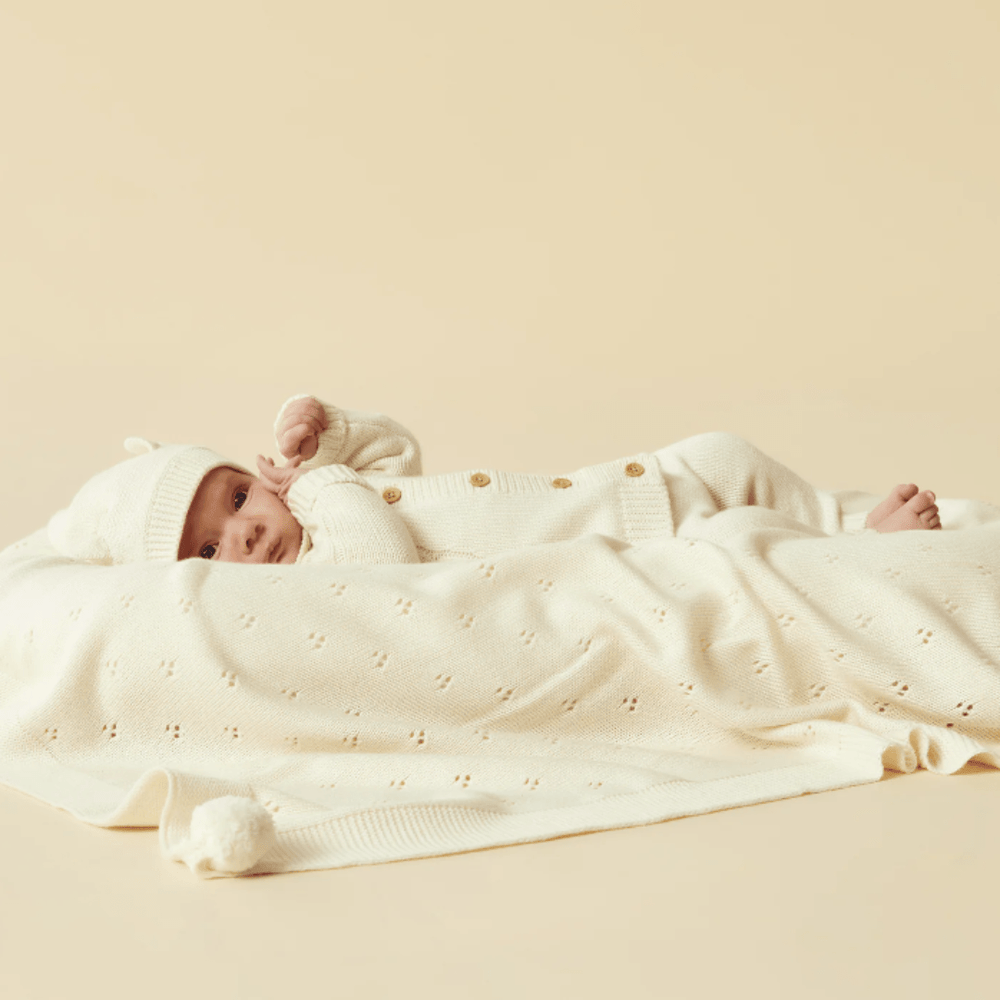 An infant dressed in a white knitted outfit and hat lying on a Wilson & Frenchy Luxuriously Soft Knitted Pointelle Baby Blanket.