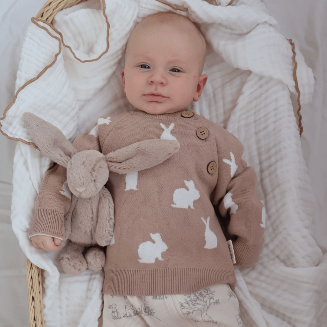 An infant wearing an Aster & Oak Organic Knit Bunny Jumper holds a plush rabbit toy while lying in a basket with a white knit blanket.