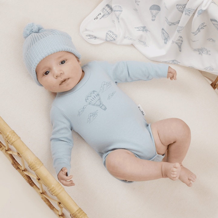 A baby wearing a blue onesie and an ethically and sustainably made Aster & Oak organic cotton beanie lies comfortably on a cream-colored surface, with a patterned blanket nearby.