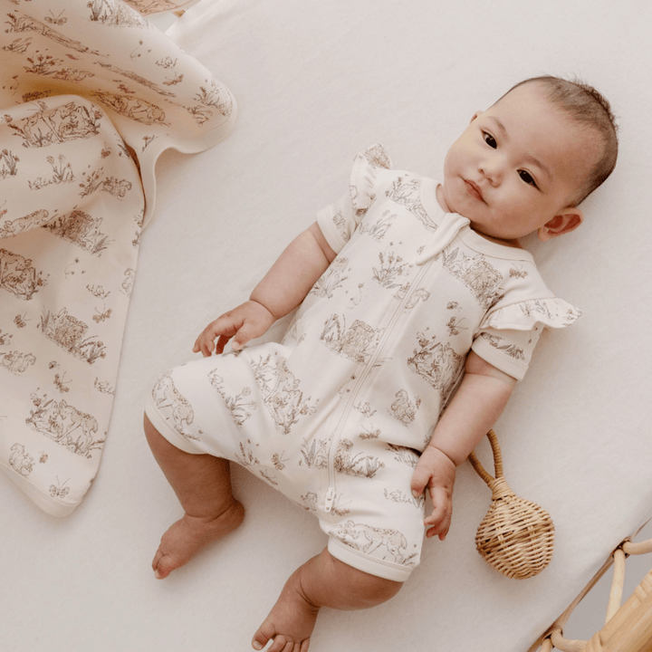 An Aster & Oak Organic Cotton Baby Swaddle Wrap is gently wrapped around a hand-illustrated print baby lying on a bed.