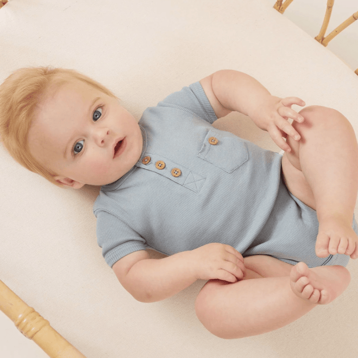 A baby with blue eyes lying down and looking upwards, dressed in an Aster & Oak Organic Rib Henley Onesie.
