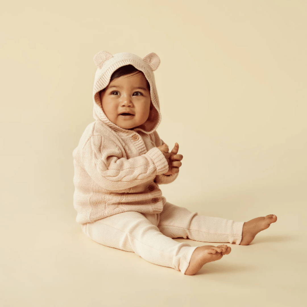Baby in a pink, Wilson & Frenchy Cable Knit Hooded Jacket with bear ears on a beige background.