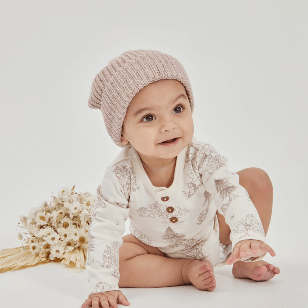 Baby in Aster & Oak Organic Henley Long-Sleeved Onesie and a knitted hat sitting with flowers nearby.
