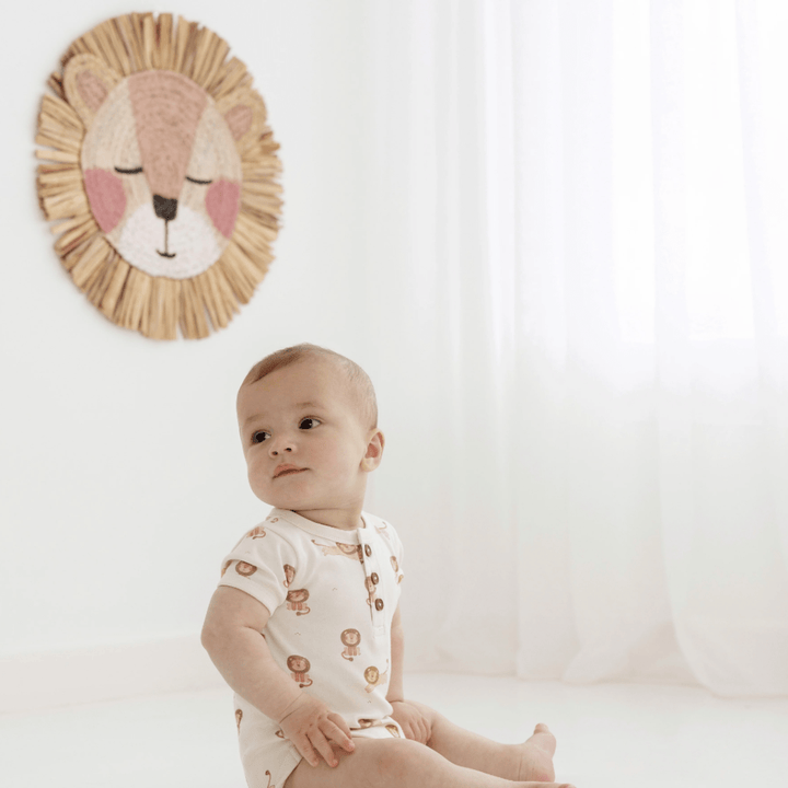 A baby in an Aster & Oak Organic Cotton AOP Henley Onesie sits on the floor, looking to the side, with a wall-mounted lion decoration in the background.