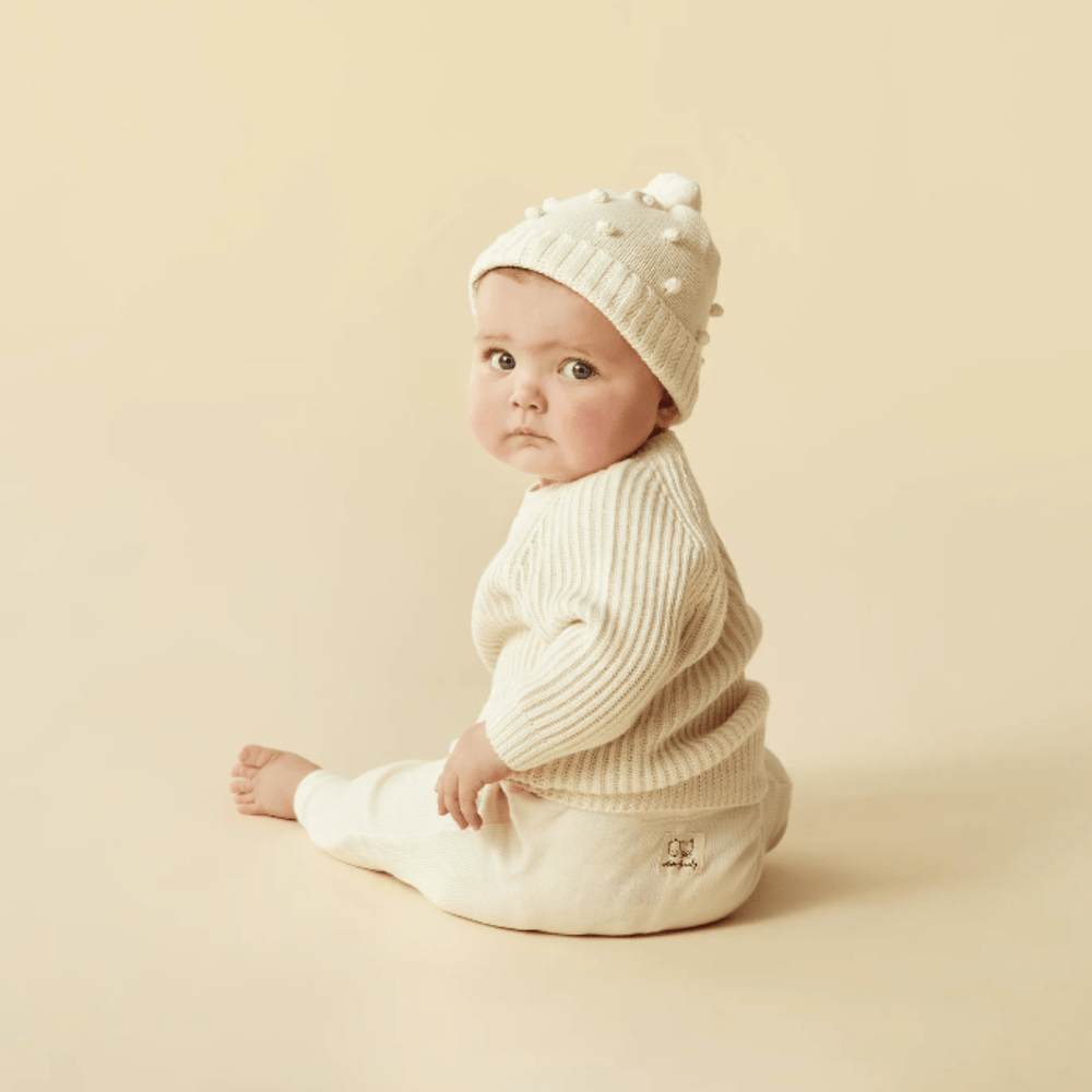 A baby in a white outfit and Wilson & Frenchy Knitted Bauble Hat sits against a beige background, looking to the side with a thoughtful expression.