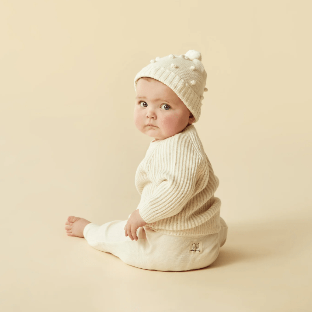A baby in a Wilson & Frenchy Knitted Ribbed Jumper and hat sitting against a beige background.