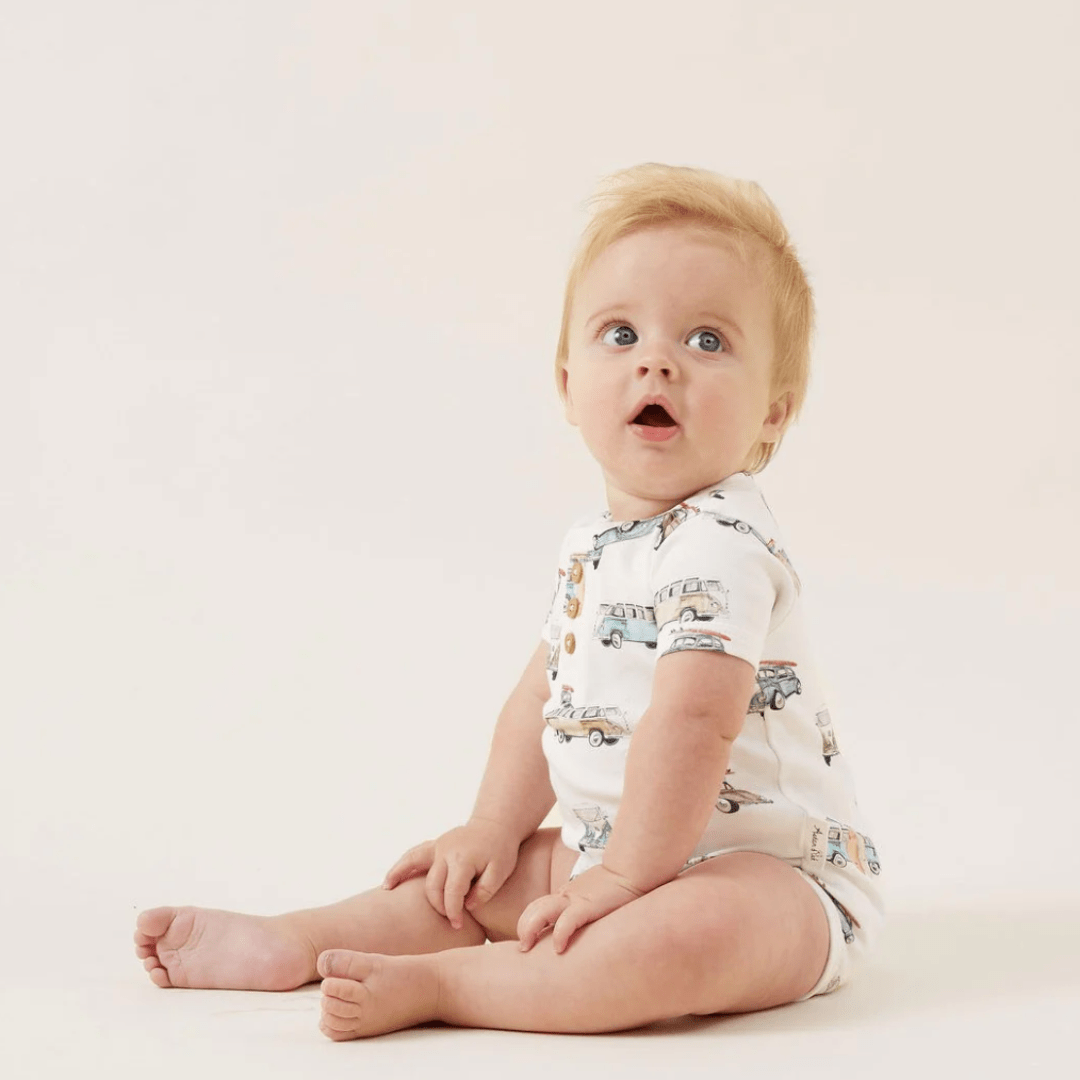Sentence with replaced product: Infant with a surprised expression wearing an Aster & Oak Organic Cotton AOP Henley Onesie - LUCKY LAST - LION - 0-3 MONTHS ONLY.