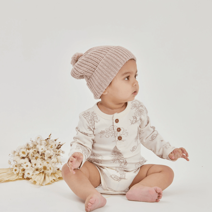 A baby wearing a beige knitted hat and an Aster & Oak Organic Henley Long-Sleeved Onesie with botanical prints sits on a white surface next to a bouquet of dried flowers.