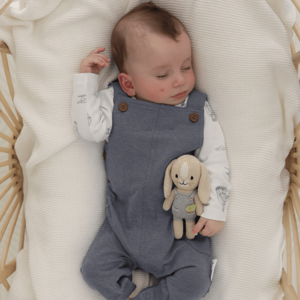 Infant sleeping peacefully in a basket with a plush bunny toy, wearing Aster & Oak Organic Dark Chambray Overalls.