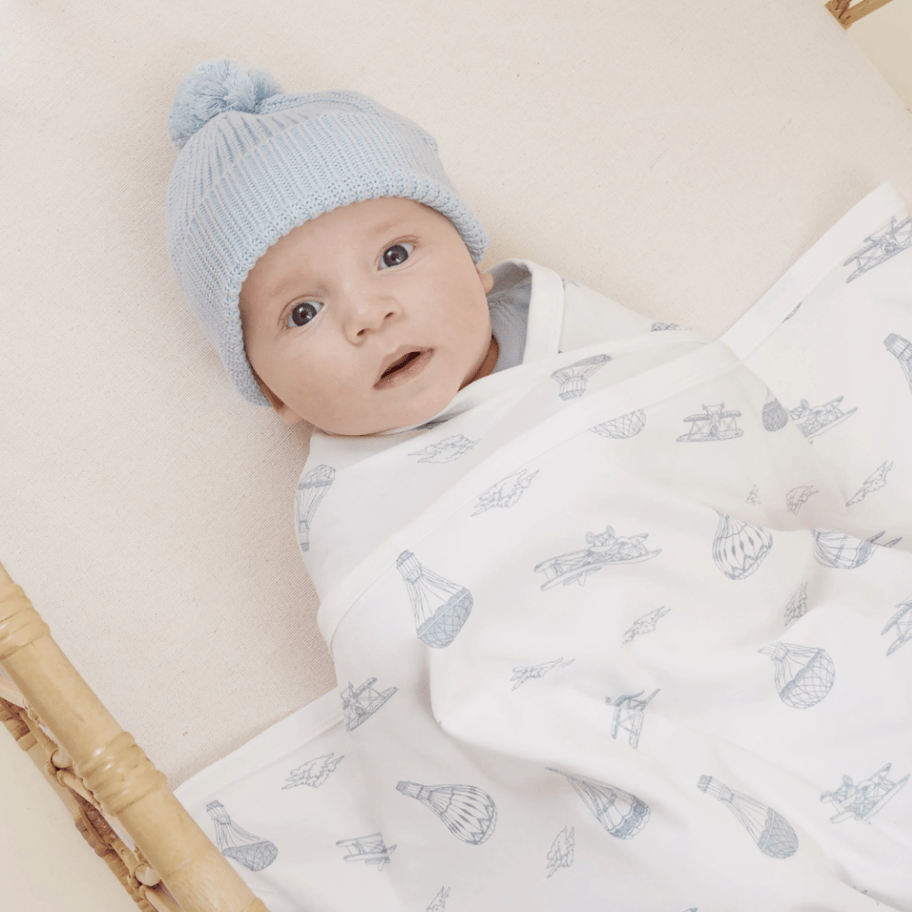 A baby wearing a blue Aster & Oak Organic Knit Beanie is lying down and wrapped in a blanket with airplane patterns.
