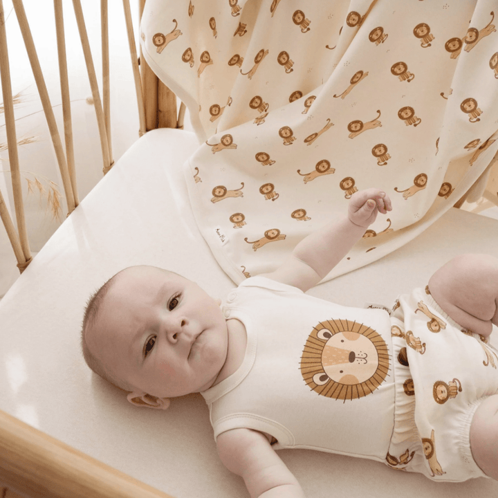 A baby swaddled in an Aster & Oak Organic Cotton Baby Swaddle Wrap, peacefully resting in a wooden crib adorned with a hand-illustrated lion print.