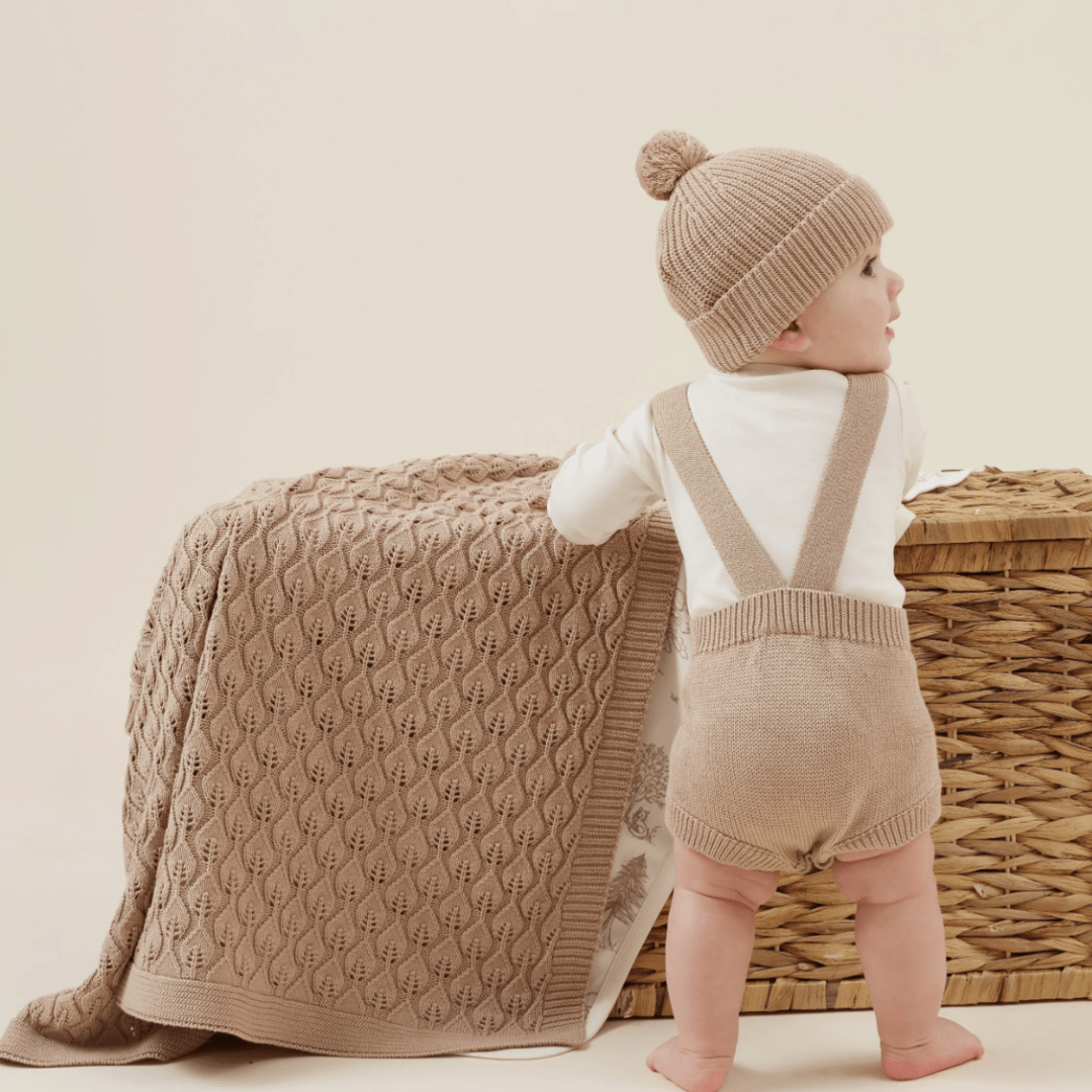 Toddler in an Aster & Oak organic knit beanie and suspenders standing beside a woven basket with a blanket draped over it.