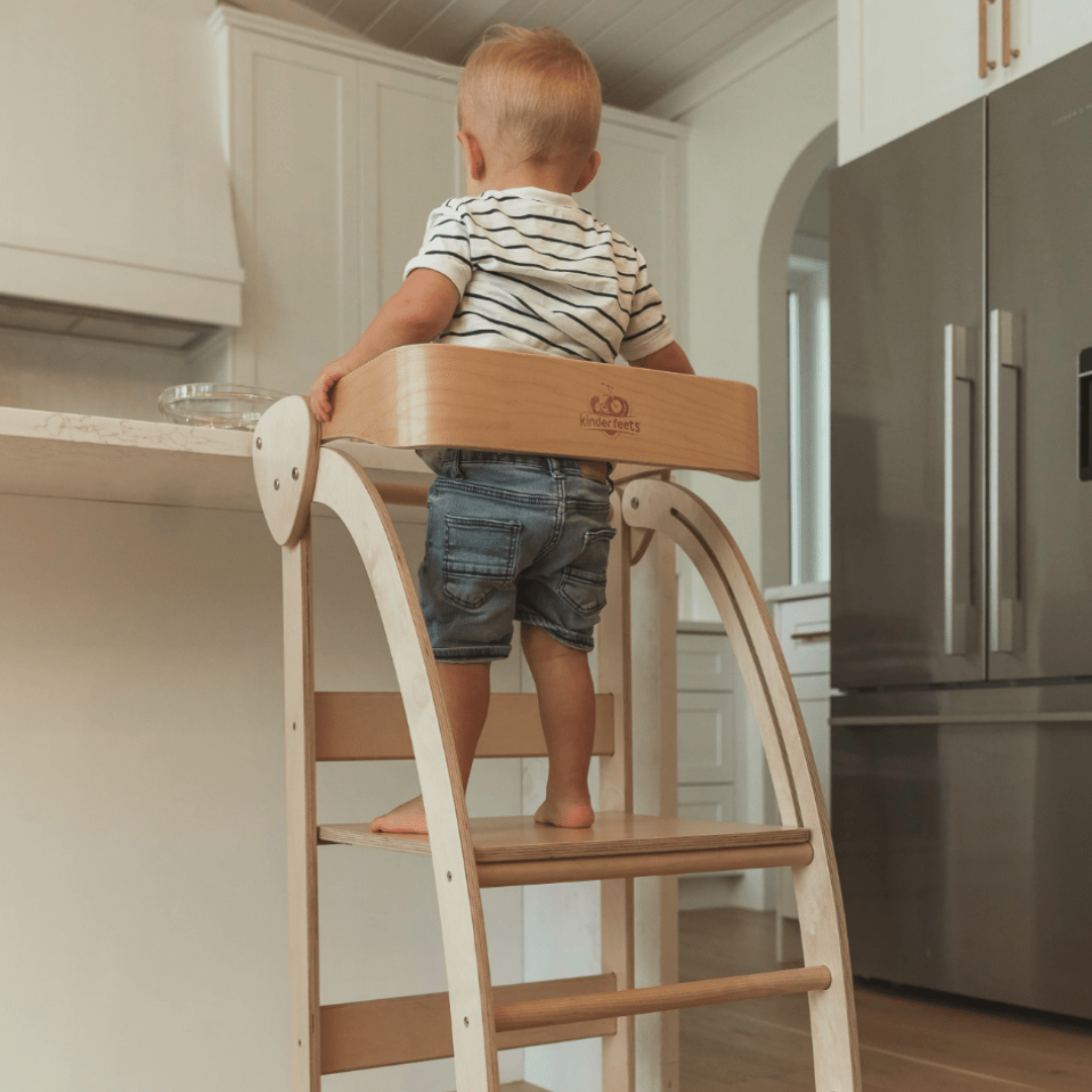 A versatile toddler engages in imaginative play while standing on a Kinderfeets Pikler Observation Tower in a kitchen.