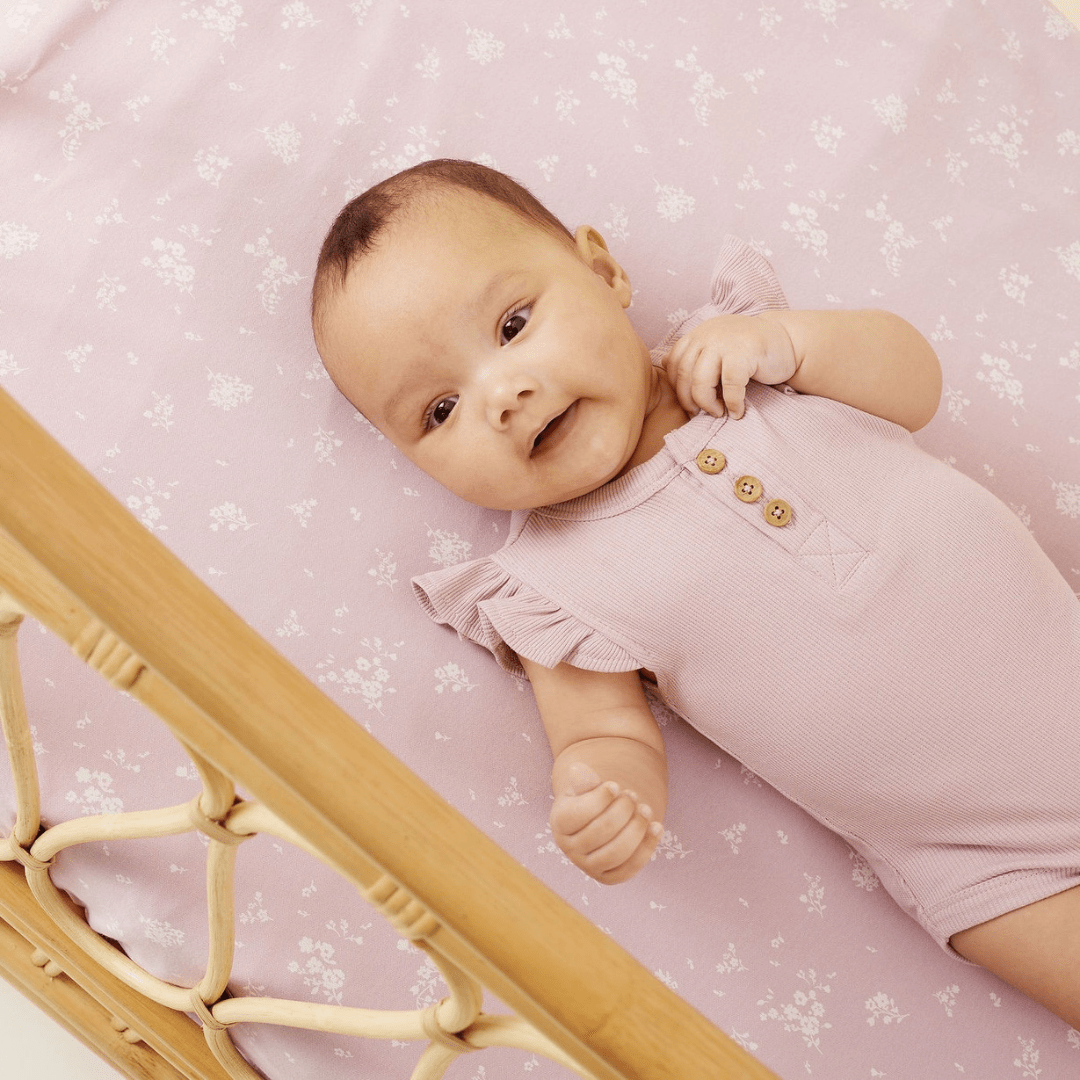 A baby is laying in a crib with a Aster & Oak Organic Cotton Dawn Pink Rib Flutter Onesie.