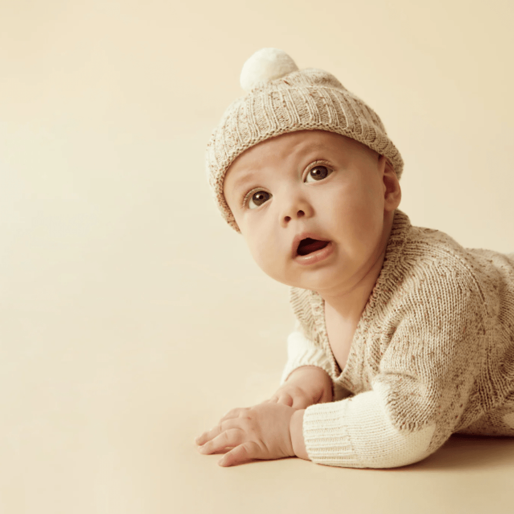 A baby wearing a Wilson & Frenchy Fleck Knitted Jacquard Jumper lying on a beige surface, looking up with a curious expression.