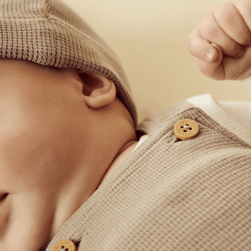 Close-up of a sleeping infant dressed in Wilson & Frenchy Organic Waffle Overalls with adjustable straps and a knitted cap.