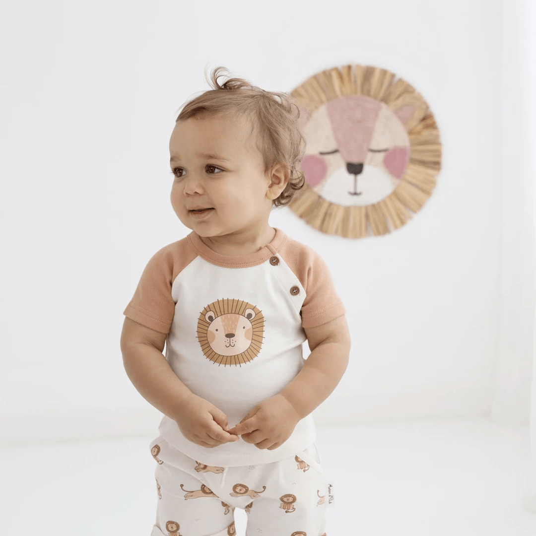 A baby is standing in front of a wall with a lion on it wearing the Aster & Oak Organic Cotton Lion Print Top made of organic cotton.