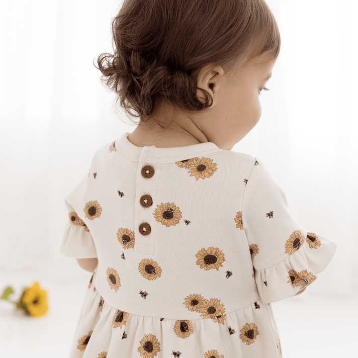 Back view of brown-haired toddler wearing dress, showing coconut buttons at the back neckline, ruffle sleeves, and beautiful hand-drawn sunflower print