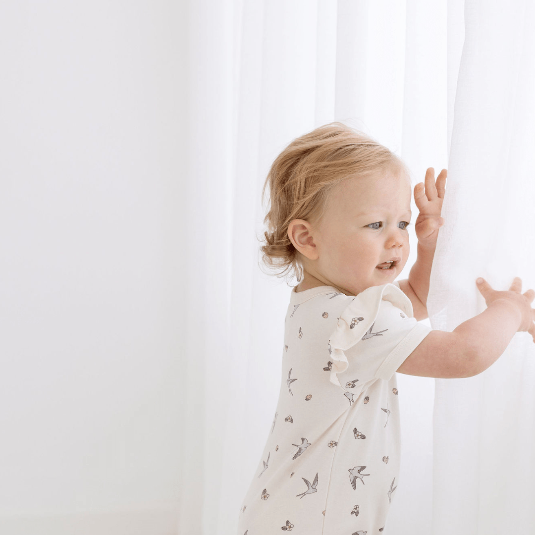 Aster & Oak, an organic cotton flutter sleeve zip romper, is reaching out of a white curtain.