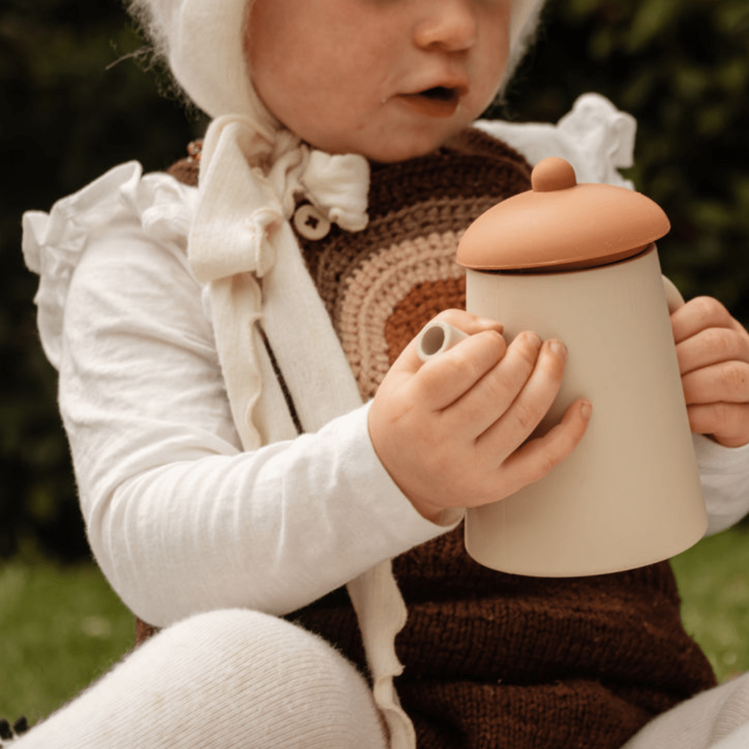 A little girl in a hat engaging in imaginative play with a Classical Child Silicone Tea Set, holding a mug.