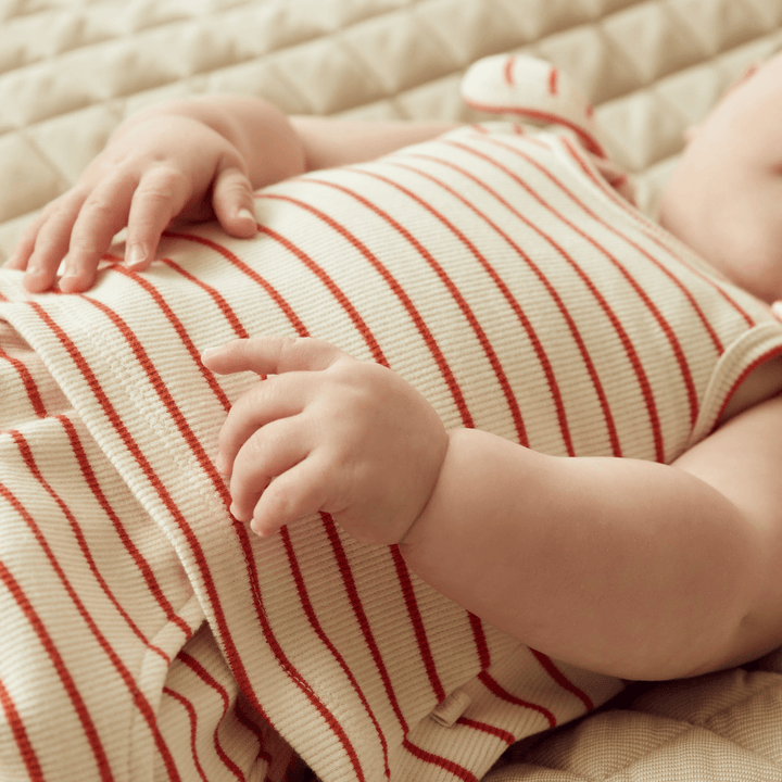 A Wilson & Frenchy baby laying on a bed wearing a Wilson & Frenchy Organic Rib Stripe Tie Singlet in red and white stripes.