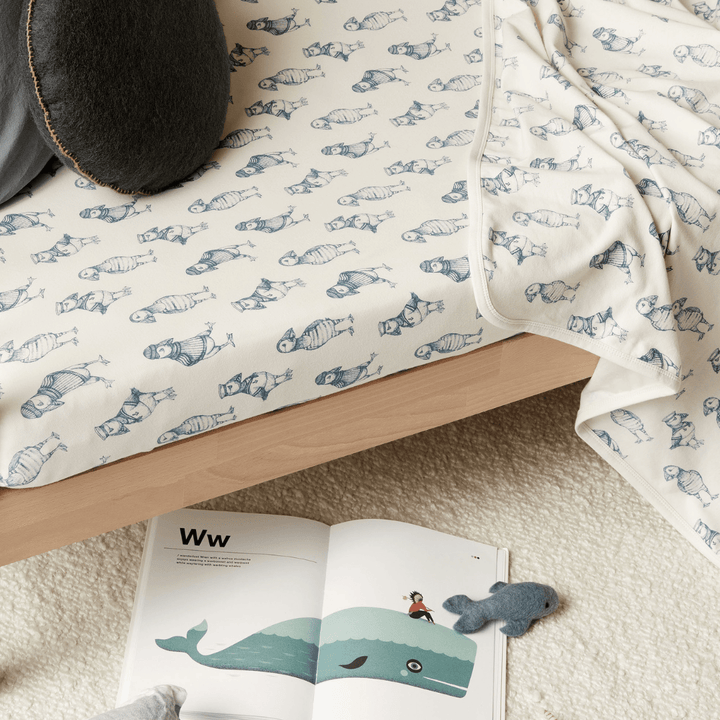 An Wilson & Frenchy Organic Cotton Cot Sheet - LUCKY LASTS - FLOAT AWAY ONLY with a baby shower present of a bed and a book on it, topped with a cozy blanket.