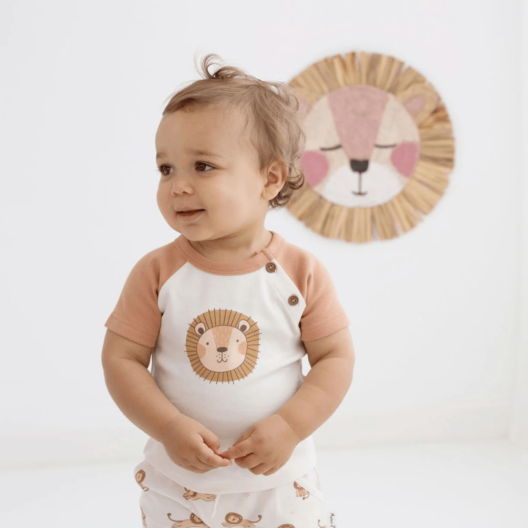 A baby, wearing Aster & Oak Organic Cotton Harem Shorts, is standing in front of a wall with a lion on it.