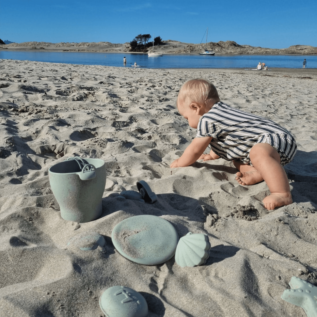 A baby is happily playing with a Classical Child silicone beach bucket set and beach moulds on a sandy beach.