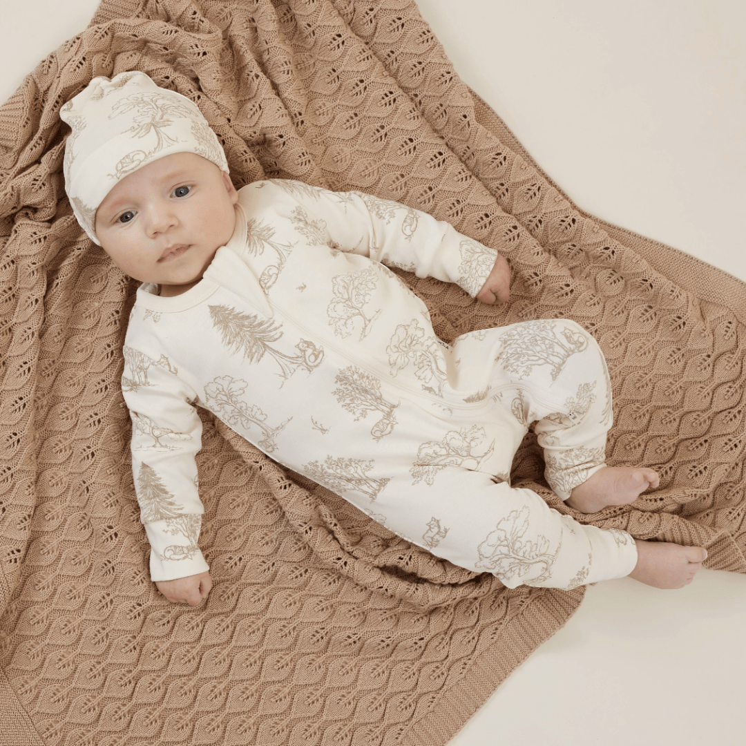 Infant dressed in a patterned onesie and an Aster & Oak Organic Knot Baby Hat lying on a knitted brown blanket.