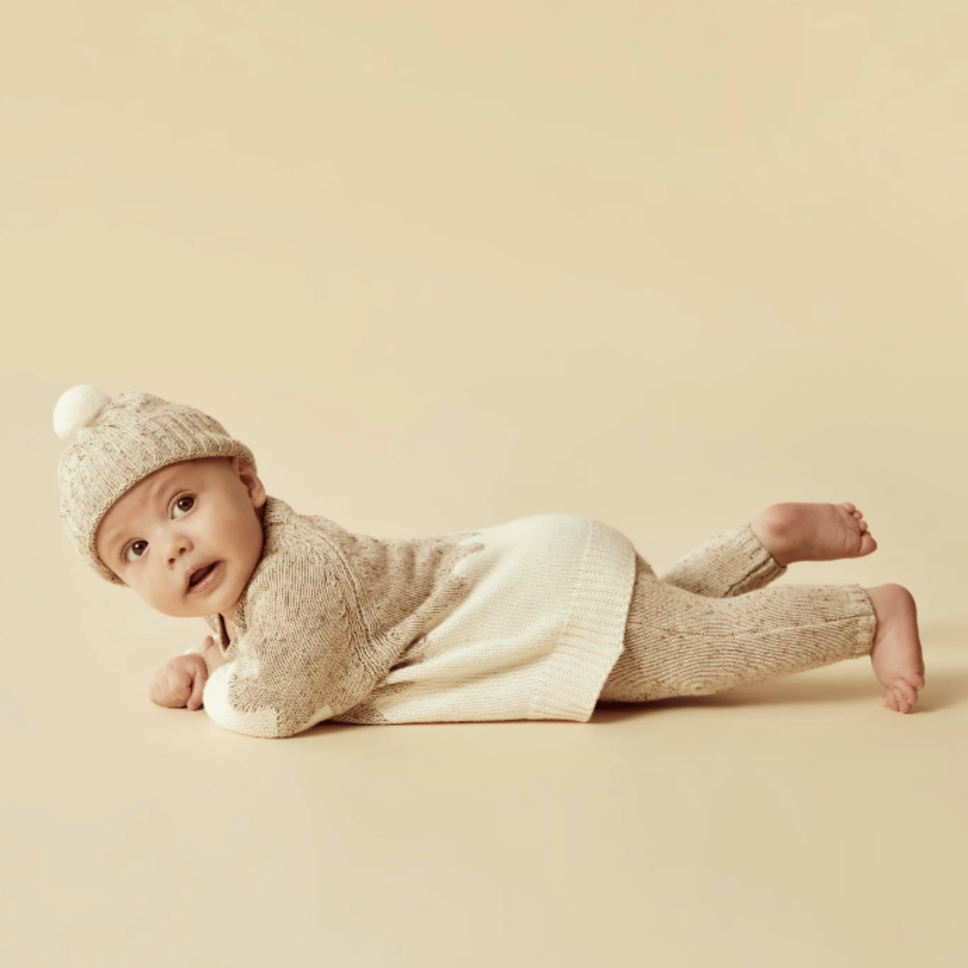 Baby lying on stomach wearing a Wilson & Frenchy Fleck Knitted Jacquard jumper and hat against a beige backdrop.