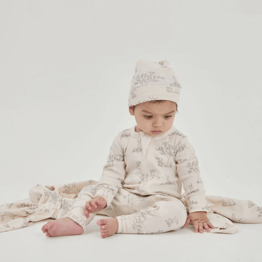 A baby dressed in an Aster & Oak Organic Bunny Luxe Rib Knot Hat and outfit sits on a plain background, looking down at their feet.