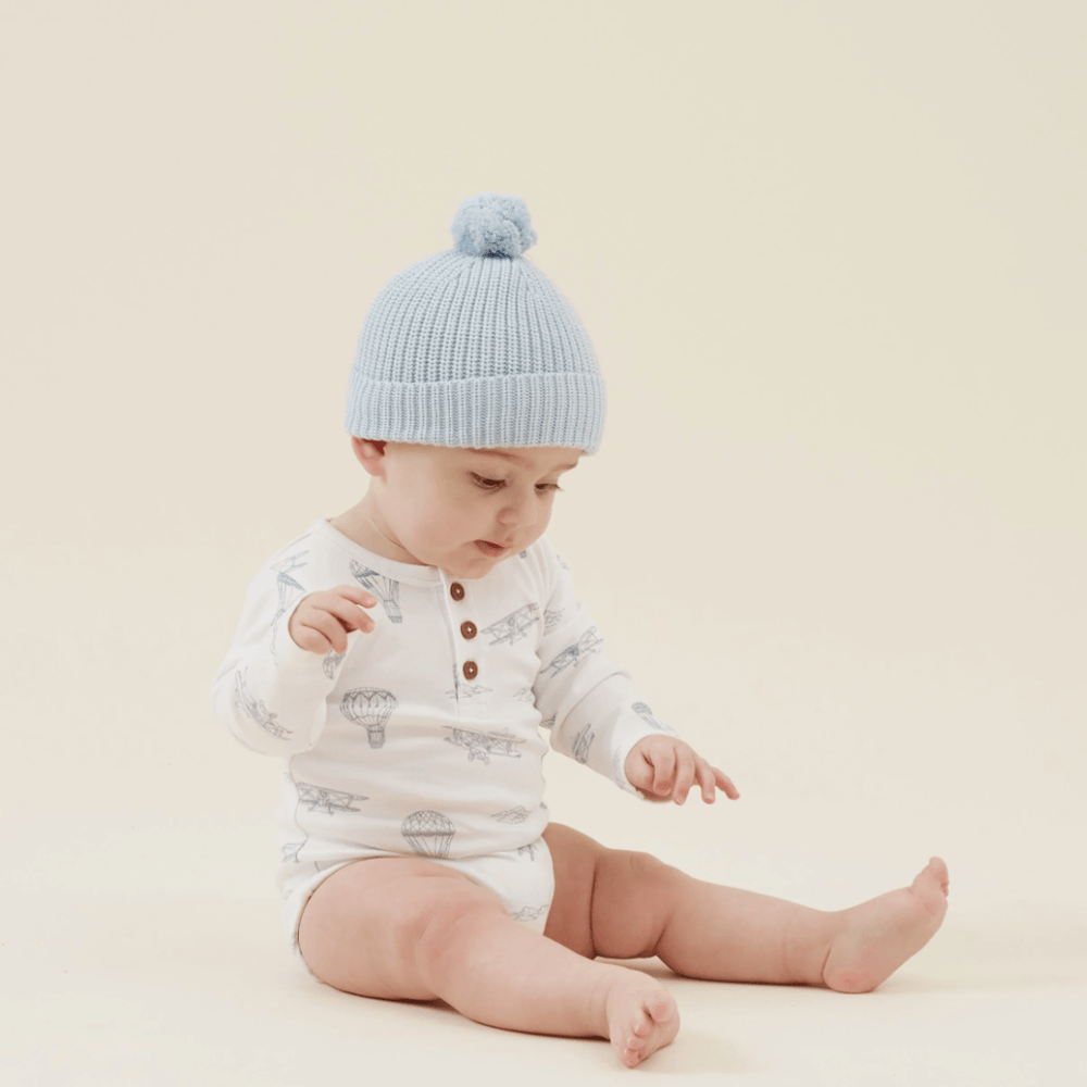 Baby sitting on a beige background wearing a pom-pom hat and an Aster & Oak Organic Henley Long-Sleeved Onesie with a hand-illustrated print.