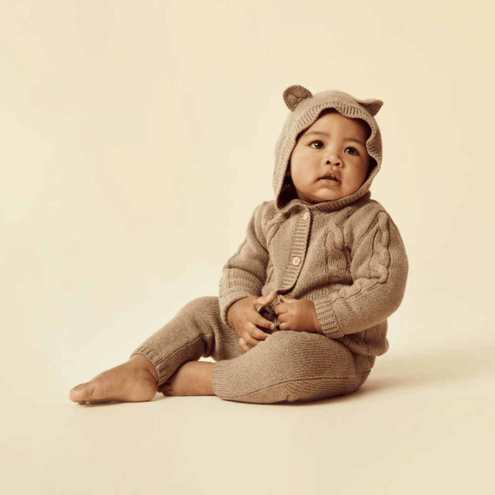 A baby wearing a Wilson & Frenchy Cable Knit Hooded Jacket sits looking thoughtful against a neutral background.