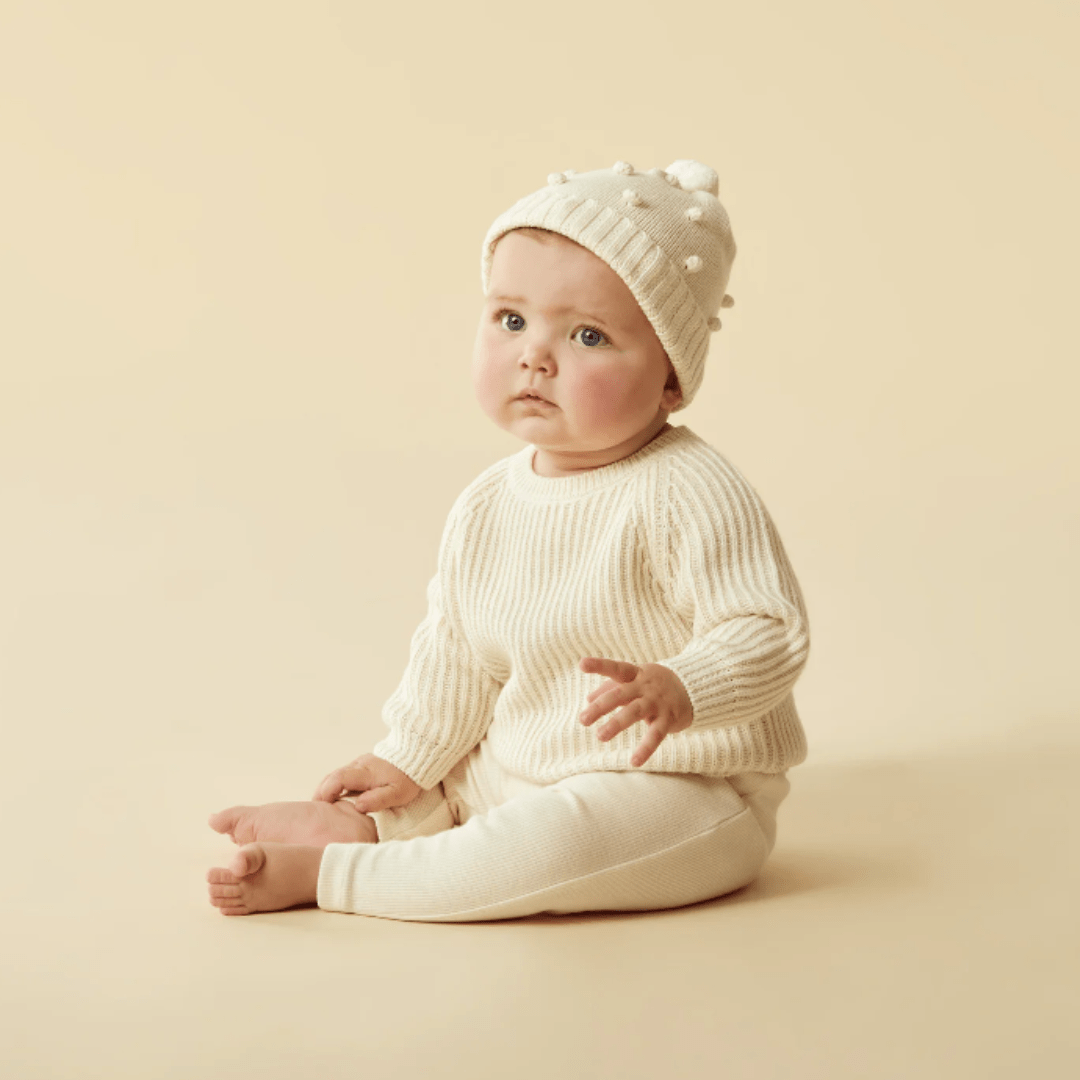 A baby wearing a cream-colored sweater and a Wilson & Frenchy Knitted Bauble Hat, sits against a beige background.